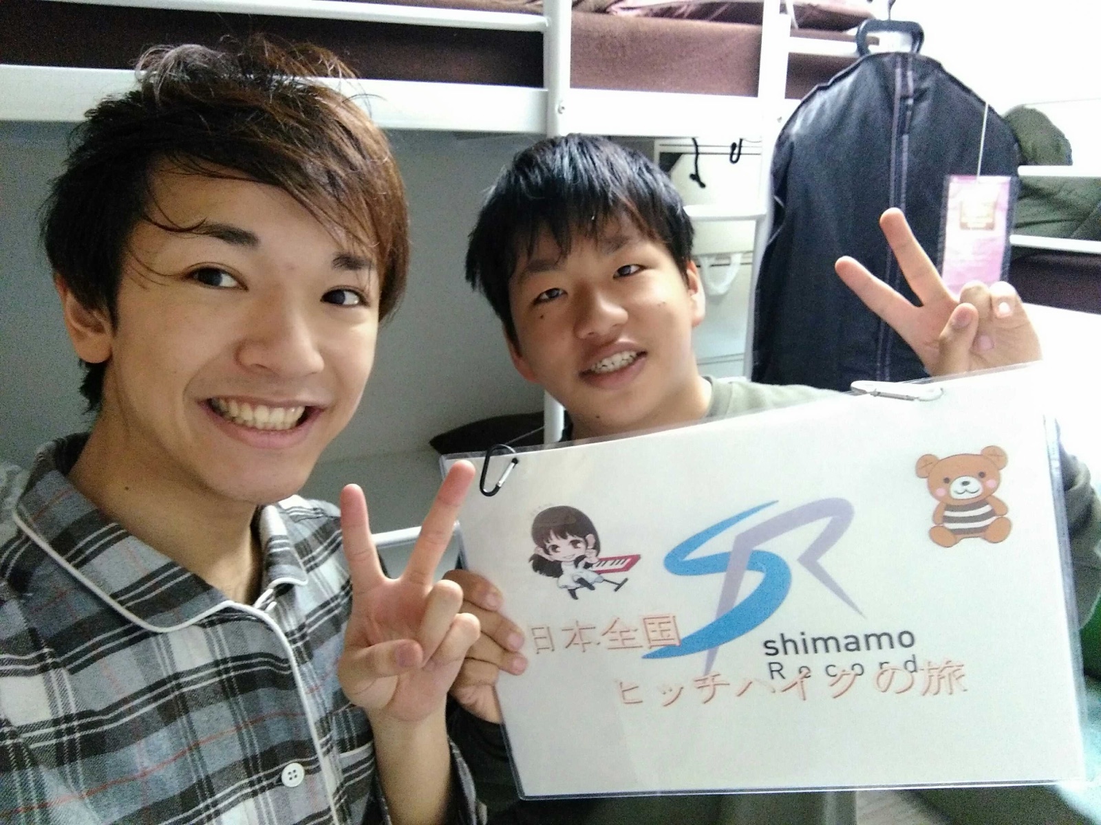 二段ベッドの前で、旅の看板を持って笑う2人